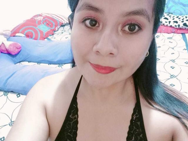 PaquitaHot, de piel clara, cabello oscuro recogido en una coleta, maquillaje de ojos rosa, lápiz labial rojo, top de encaje negro con tirantes, pequeño piercing en la nariz, sonriendo a la cámara, mostrando sus pechos..