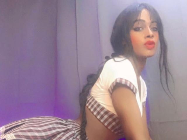 KataleyaCollins with long black hair, red lipstick, and a checkered top, leaning forward, showing her back and a small butt, wearing a plaid skirt, in a soft light.