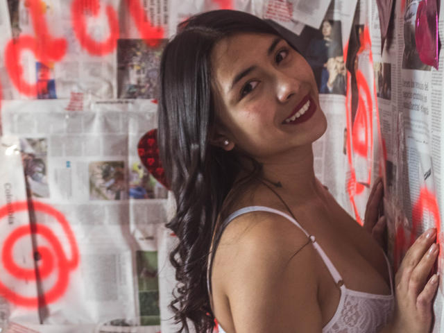 IsabellaPrincess with long black hair, smiling, wearing a white lace bra, standing against a wall of newspaper, red graffiti, and photos, showing a cute face and small tits.