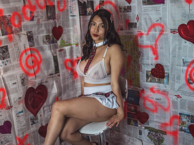 Isabella, una principessa con capelli scuri, labbra rosse, reggiseno bianco, cravatta a quadri e gonna bianca, seduta su una sedia, che mostra i seni e i glutei, con uno sfondo di giornale con cuori rossi..