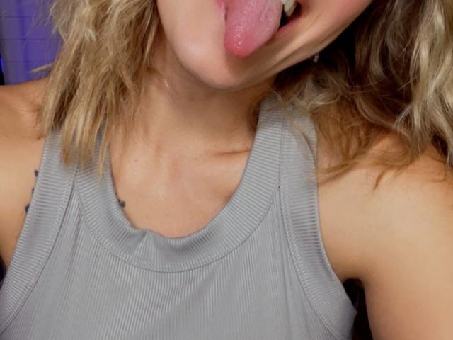 Misstyk's pink tongue is sticking out, showing her teeth, with wavy blonde hair and wearing a gray ribbed tank top, exposing her collarbone.