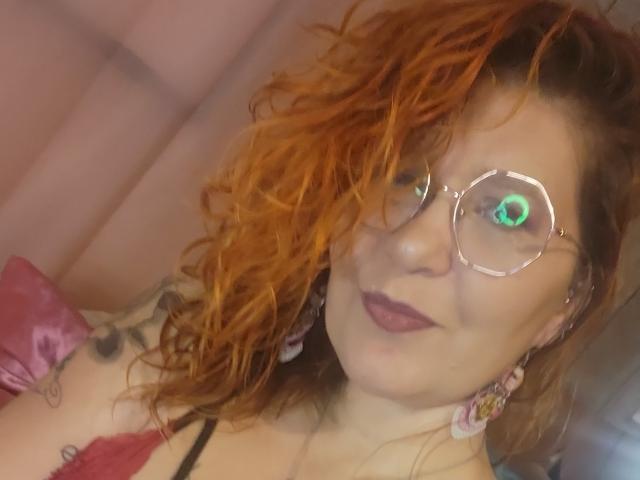 Choukette, avec ses cheveux orange bouclés, ses lunettes rondes, son rouge à lèvres rose, ses tatouages et ses boucles d'oreilles roses, montre ses seins dans un soutien-gorge rouge, et sourit à l'appareil photo..