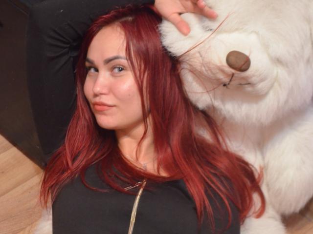 MissKaryna, une rousse avec un anneau de nez, portant une chemise noire, avec un ours en peluche blanc à sa droite, allongé sur un oreiller noir, et un sol en bois en arrière-plan..