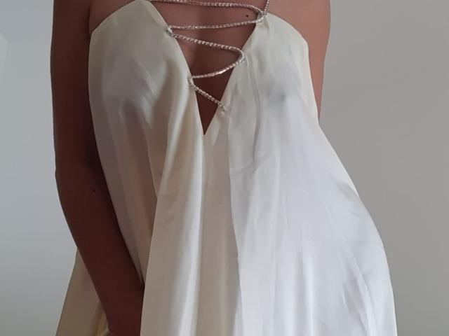 Vitche69 indossa un abito bianco in raso con un profondo scollo a V, decorato con catene d'argento che attraversano il petto. Il seno è parzialmente visibile attraverso il tessuto. Sfondo morbido e di colore chiaro..