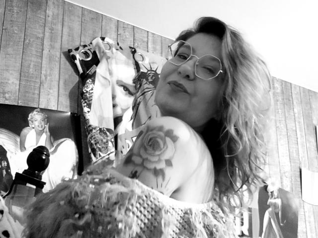 Fotografía en blanco y negro de Choukette con cabello rizado, gafas y un tatuaje de rosa en el hombro, guiñando un ojo, vistiendo un suéter suave, de pie en una habitación con carteles de Marilyn..