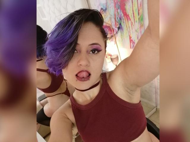 CassieJones with purple and black hair, tongue out, wearing a maroon sports bra, visible armpit, and a choker, selfie in a mirror, showing her ass in the background.