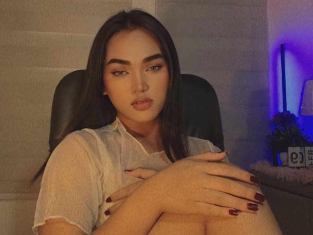 Princesa Kylie, con cabello castaño largo, cejas pobladas y uñas rojo oscuro, sentada en una silla negra, vestida con una blusa blanca transparente, con las manos sobre las rodillas, mirando a la cámara..