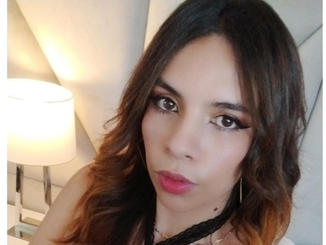 VioletaStone has medium brown skin, long wavy brown hair with orange tips, thick black eyeliner, pink lipstick, and a black lace choker, looking into the camera with pouty lips.