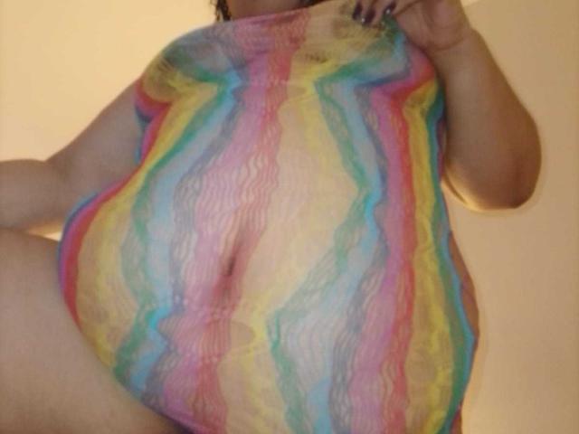 FoxyBell trägt ein durchsichtiges Oberteil mit Regenbogenstreifen, das ihre großen, runden Brüste und einen Teil ihres weichen, geschwungenen Bauches zeigt. Ihre Hand ist teilweise sichtbar..