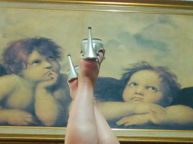 CoupleMature's legs with silver high heels, crossed in front of a painting of two cherub-faced babies with curious expressions. Babies have fluffy brown hair, looking upward.