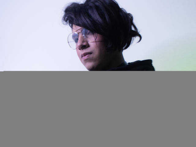 Saturni with messy black hair, glasses, and a black hoodie with red lace-up sleeves, looking slightly confused. Soft blue-green light on the right.