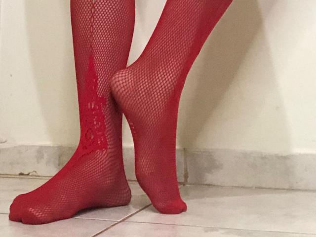 Le gambe di MarryCassol, coperte da calze a rete rosse, sono in piedi su un pavimento piastrellato, con il piede sinistro incrociato sopra quello destro, mettendo in risalto i contorni e la texture dei suoi piedi..