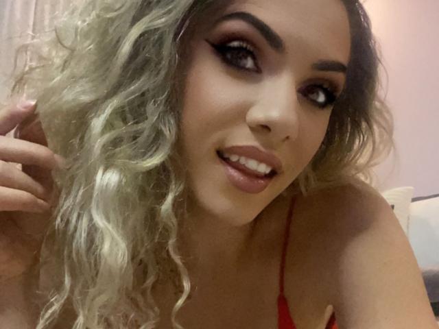 LucilleXSugar, con cabello rubio rizado, delineador de ojos oscuro y lápiz labial rojo, sonriendo con la mano cerca de su rostro, vistiendo una blusa roja, mostrando ligeramente sus senos..