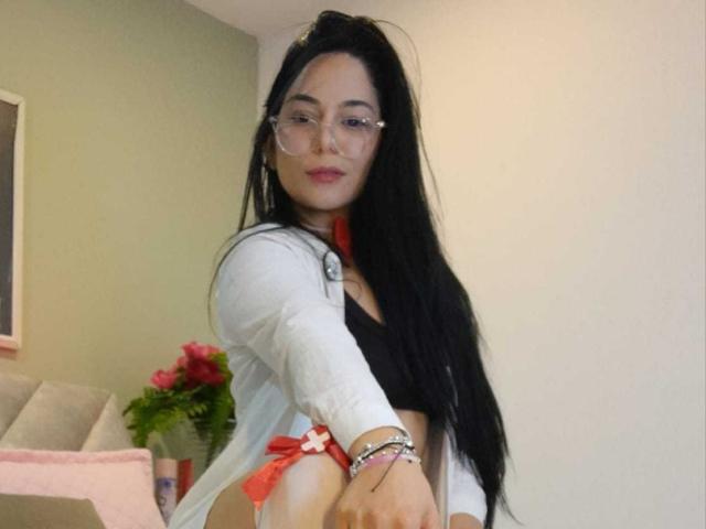 BiancaScott with long black hair, glasses, white cardigan, black bra, red bow on her side, looking at camera, smiling, hands on hips, red flowers in background.