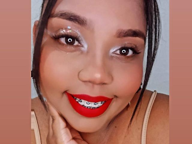 SarahSmity tem olhos castanhos brilhantes, batom vermelho e um leve sorriso, com cabelo preto preso em um rabo de cavalo e uma mão tocando sua bochecha. Ela está vestindo uma blusa bege..