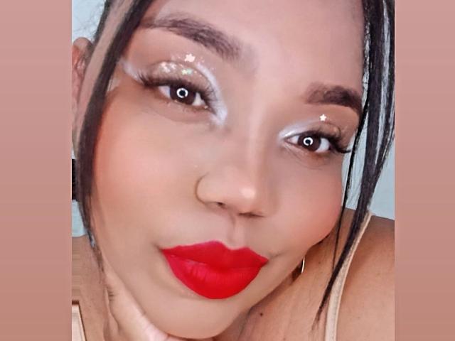 SarahSmity's face with shiny white eye makeup, red lipstick, and black hair in a side part, looking directly at the camera with a subtle smile. Boob outline visible.