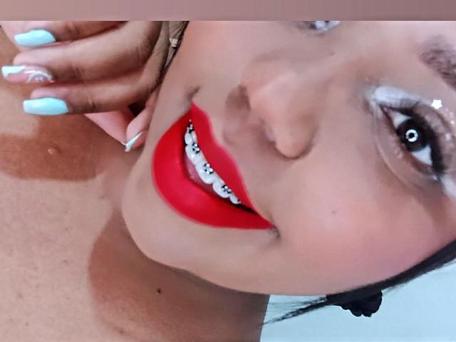 El rostro de SarahSmity, con lápiz labial rojo, dientes blancos, esmalte de uñas azul y piel morena. Su mano, con uñas azules, toca su mejilla. Un clip de oreja negro en el lado derecho..