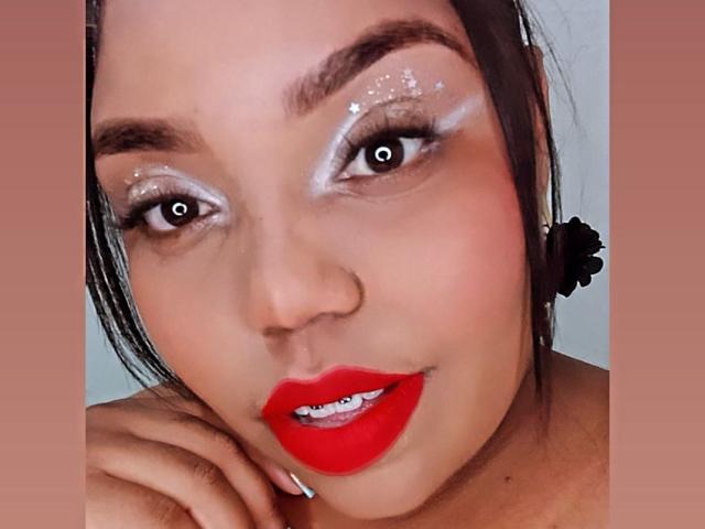 SarahSmity has shiny silver eyeshadow, sparkling glitter, red lipstick, and black eyebrows. Her brown eyes are wide, and she's smiling with her hand near her face.