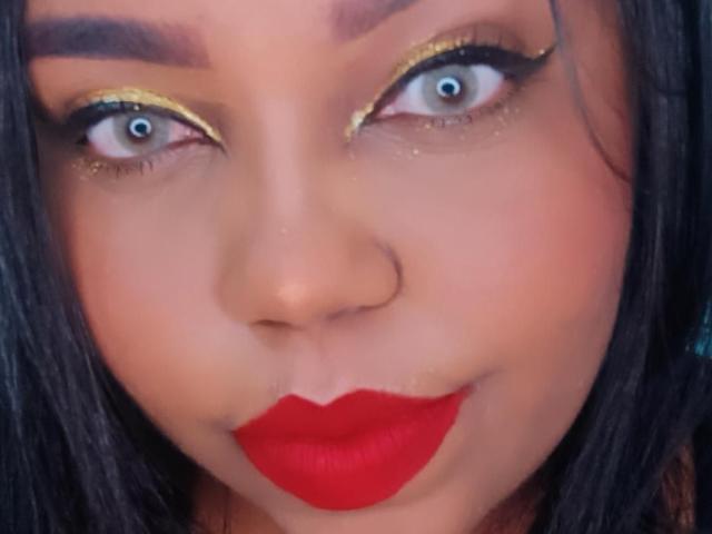 SarahSmity's face with bright blue eyes, golden eyeliner, and red lipstick, showing a slightly surprised or playful expression, with dark brown hair and a small nose.