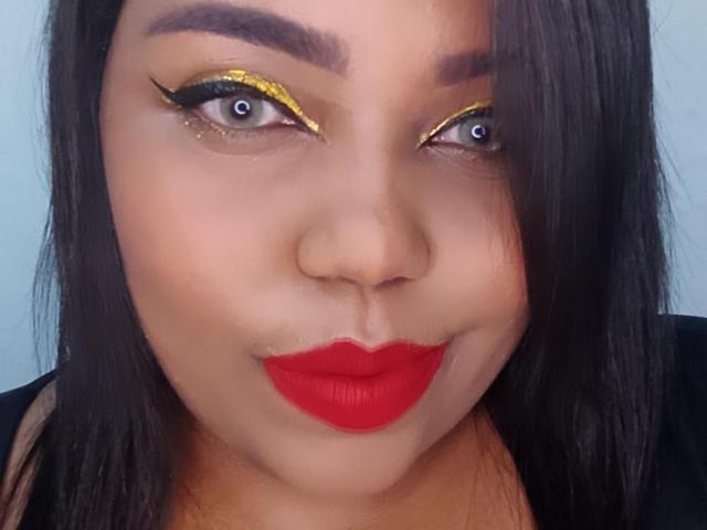 SarahSmity tem um delineador amarelo brilhante, sobrancelhas pretas grossas, batom vermelho e olhos castanhos com um leve sorriso. Seu cabelo preto é liso e ela está vestindo uma blusa preta..
