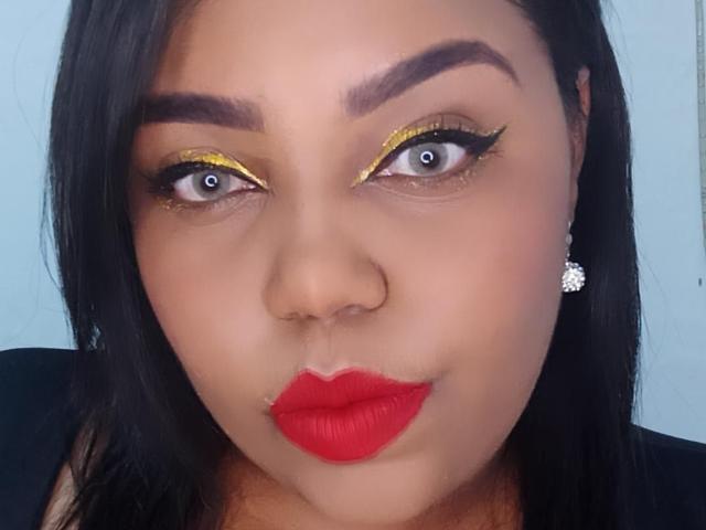 Sarah Smity hat gelben Eyeliner, rote Lippen, dunkle Augenbrauen und blaue Augen, trägt Diamant-Ohrringe, hat schwarzes Haar und ein Nasenpiercing und blickt direkt in die Kamera..
