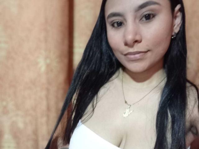 PaulaBigBoobss with long black hair, brown eyes, and a subtle smile, wearing a white strapless top, gold necklace, and earrings, showing her tits against a soft orange background.