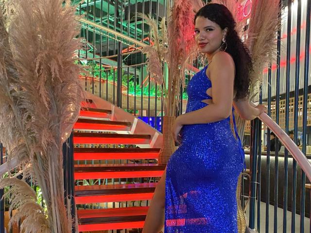TerraFoxy stands on a red staircase, wearing a blue, sparkly dress that hugs her ass and shows a thigh-high slit, with puffy grasses on either side.