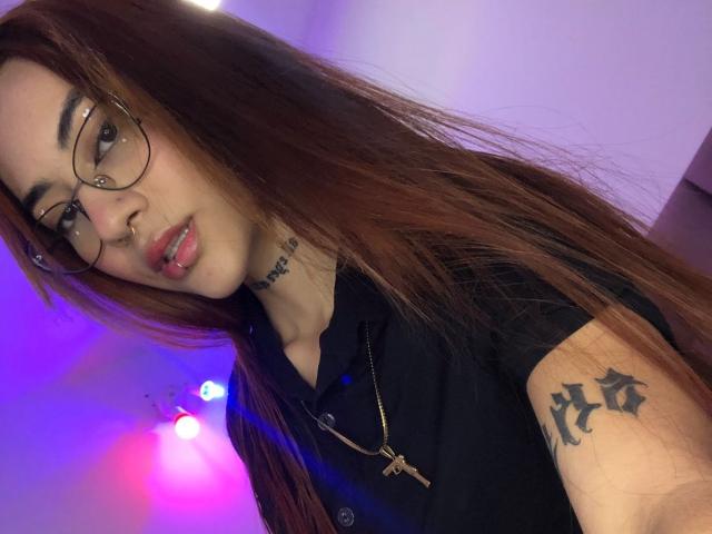 CieloSmith mit langen, braunen Haaren, Brille, schwarzem Hemd, Nasenpiercing, Lippenpiercing, goldener Halskette, Tattoo am rechten Arm, violett-blauer Beleuchtung und einem selbstbewussten, leicht verführerischen Ausdruck..