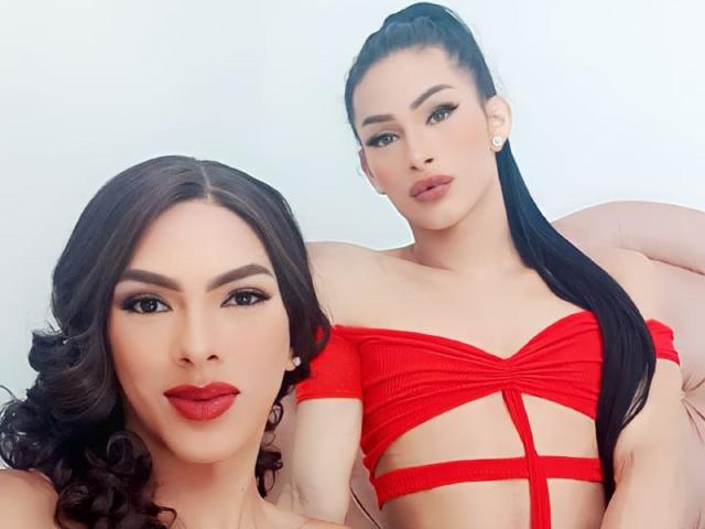 BelivianCoupleTs with curly brown hair and red lipstick, and BelivianCoupleTs with long black hair in a ponytail, wearing a red strapless top, showing their tits.