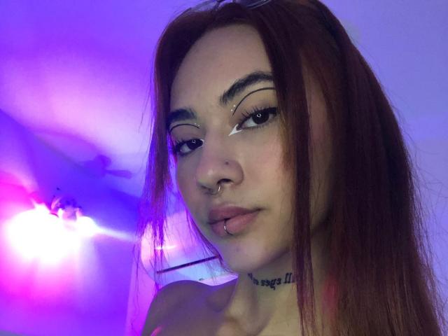 CieloSmith ha i capelli castani, un piercing al naso e un piercing al labbro. Sta guardando la fotocamera, con una luce al neon viola e blu sullo sfondo. Il suo seno non è visibile..