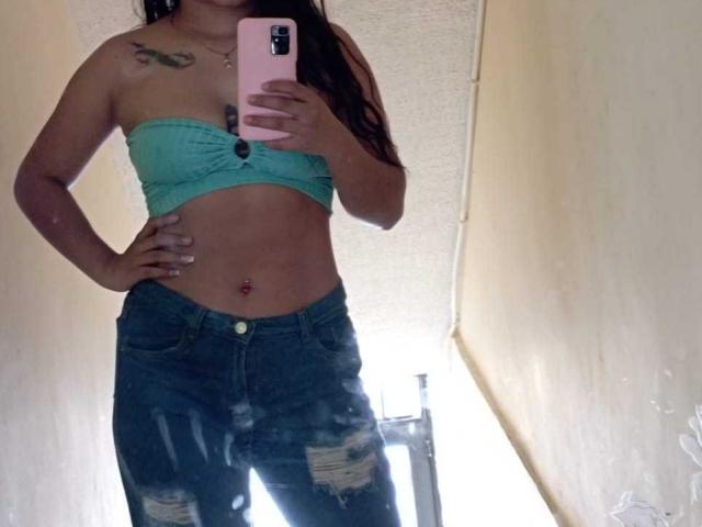 Kratos e Samira, con la pelle abbronzata, un top a tubino color turchese e jeans strappati, che mostrano un addome piatto, un piercing all'ombelico e un tatuaggio sulla spalla destra, stanno scattando un selfie allo specchio..