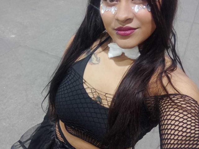 KratosYSamira, con cabello negro largo, lleva puesto un top de rejilla, mangas de malla negra, un collar blanco y lápiz labial rosa. Tiene brillo en las mejillas y un tatuaje sobre los pezones..