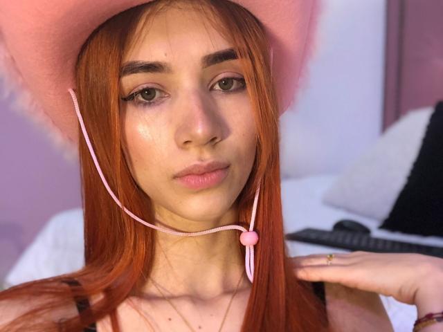 Greta Harris, met rood haar, draagt een roze hoed en een roze headset met een hart. Ze heeft een lichte huid, groene ogen en subtiele make-up, en kijkt naar de camera..