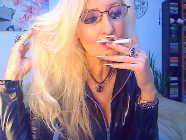 DominatrixChris with long blonde hair, glasses, and a black leather jacket, sucking on a cigarette with long white nails, wearing a purple pendant and rings, smiling.