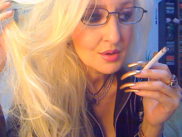 Blonde DominatrixChris with long hair, glasses, and gold nails, holding a cigarette, wearing a black zip-up top, and a silver necklace, looking down with a slight smirk.