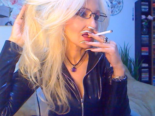 DominatrixChris with long blonde hair, wearing glasses, black leather jacket, holding a cigarette, purple ring necklace, and ring, fingers with white nail polish, sexy and seductive.