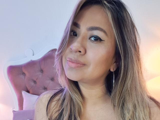 ValeriaFranco tem olhos castanhos, cabelo castanho claro e um sorriso discreto com lábios rosados. Ela está usando pequenos brincos de argola e parece adorável, com uma cabeceira rosa e texturizada atrás dela..