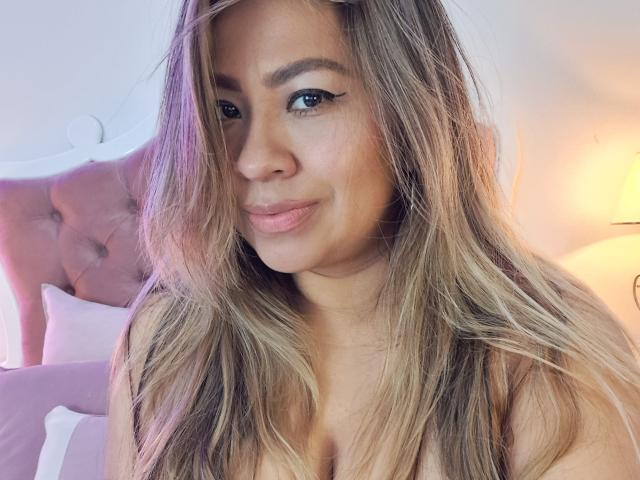 Valeria Franco, met lang blond haar, subtiele eyeliner en een zachte glimlach, laat haar borsten lichtjes zien, terwijl ze in een zacht verlichte slaapkamer zit..