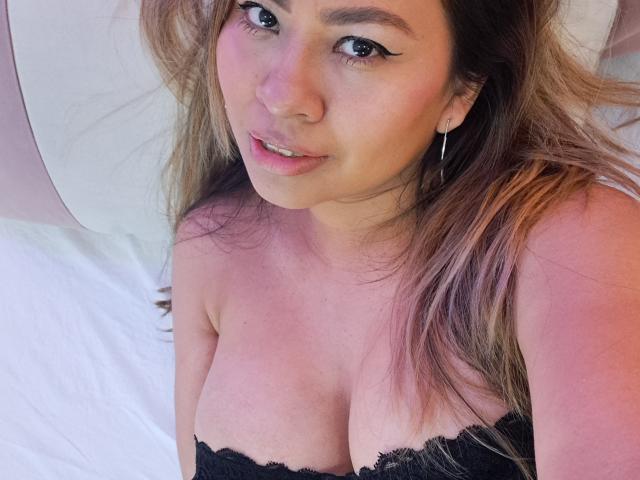 ValeriaFranco, avec ses longs cheveux bruns, portant de la lingerie en dentelle noire, montrant ses seins volumineux, regardant vers le haut avec du crayon noir pour les yeux, de petites boucles d'oreilles et un léger sourire..