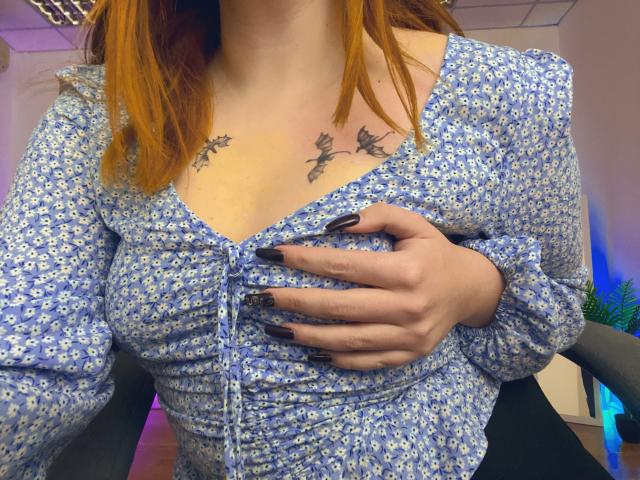 Una donna dai capelli rossi, con un abito floreale blu e unghie nere, che si tiene i seni, mostrando tatuaggi a forma di farfalla sopra il petto. Ambiente d'ufficio, con luci a soffitto visibili..