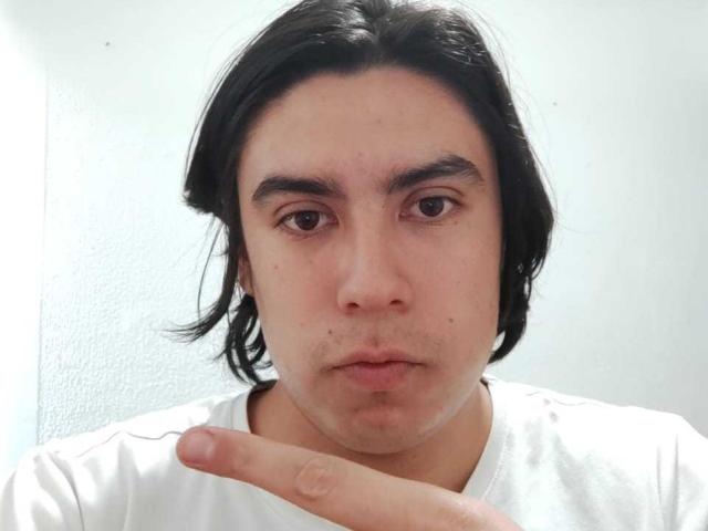 Zoordon has medium-length black hair, brown eyes, and a neutral expression. He's pointing at his chin with a finger, wearing a white shirt.