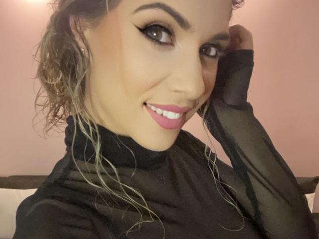 LucilleXSugar a les cheveux blonds bouclés, porte un haut noir transparent, un trait d'eye-liner noir épais, du rouge à lèvres rose et elle a un sourire. Elle touche sa tête avec une main, mettant ainsi en valeur ses seins..