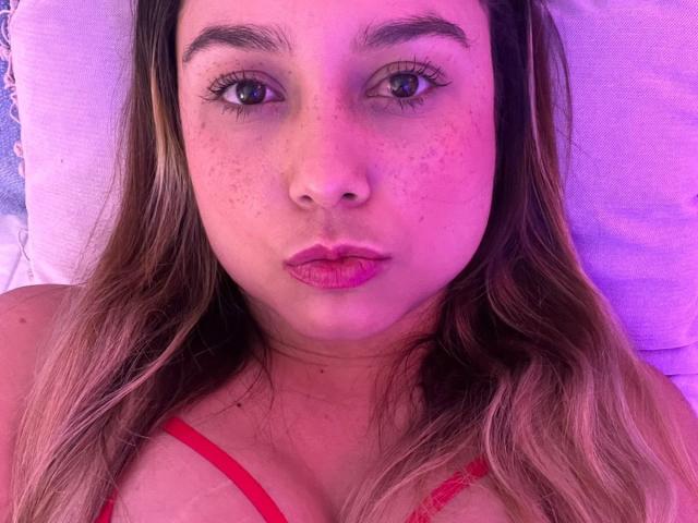 AmayaJhons with pink-tinted skin, brown eyes, and freckles, wearing a red bra, lying down, looking at the camera with pink lips, long brown hair, and a serious expression.