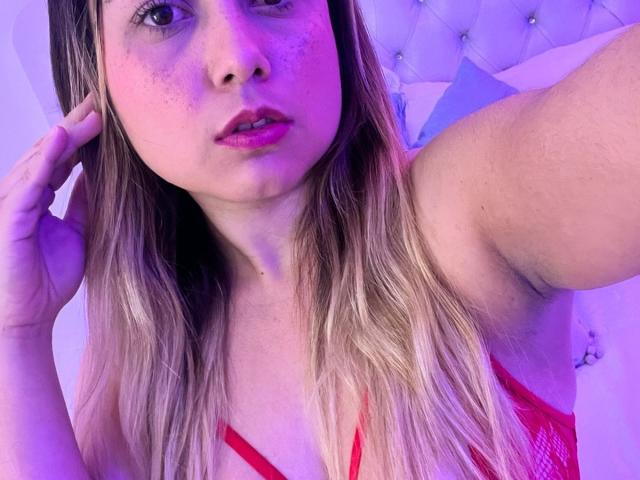 AmayaJhons, avec de longs cheveux blonds, du rouge à lèvres rose et de la lingerie rouge, tenant sa tête, montrant ses seins, sous un éclairage violet et bleu, regardant la caméra..