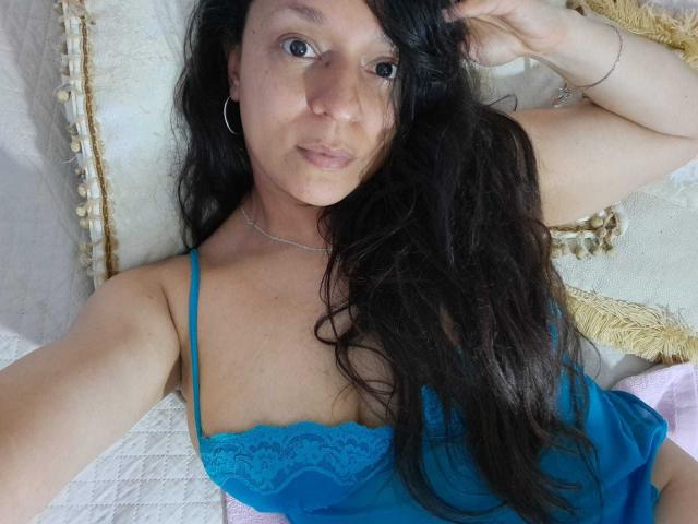 Angelonix with long black hair, blue lace lingerie, hoop earrings, and a silver necklace, lying on a bed, hand in hair, looking at camera, slightly smiling, visible tits.