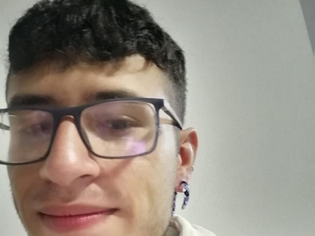 KeithThops tem cabelo curto e escuro, usa óculos e tem um sorriso discreto, com um pequeno piercing na orelha esquerda. Sua pele é clara e ele está vestindo uma camisa branca..