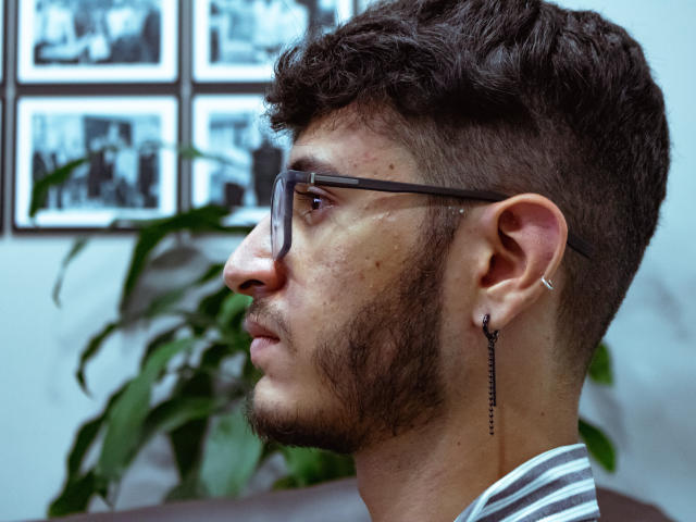 KeithThops a une barbe brune bouclée, des lunettes, et un boucle d'oreille noir avec une chaîne, et il regarde sur le côté avec une expression sérieuse. Sa peau est légèrement constellée de taches de rousseur..