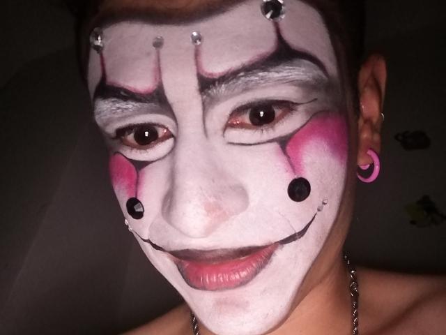 KeithThops has white face paint with black and pink accents, pierced ears with pink plugs, and a chain necklace, smiling with dark eyes and short brown hair.