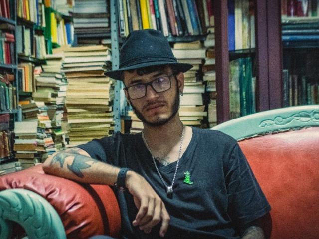 Keith Thops, usando óculos, com um chapéu preto e um braço tatuado, apoiado em um sofá de couro vermelho, vestindo uma camiseta preta e um colar, em um quarto cheio de livros..