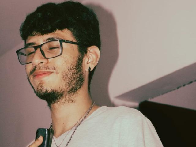 KeithThops, avec ses cheveux bouclés foncés, ses lunettes, sa barbe et son boucle d'oreille, souriant les yeux fermés, tenant un téléphone, portant un t-shirt blanc et un collier en chaîne..